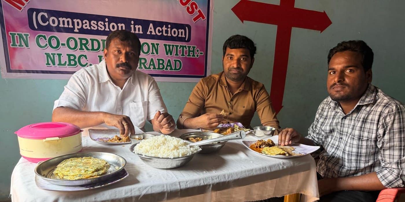 MERCE Charitable Trust leaders sharing a meal with community members — Compassion in Action banner visible